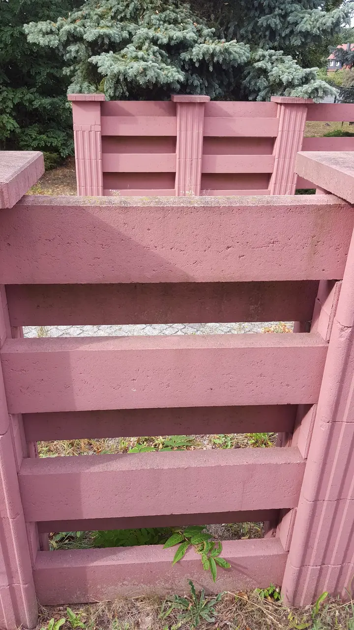 Blick in eine rote Einfassung aus Vablo Pfosten und Betonriegeln, im Hintergrund Nadelbäume