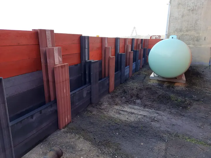 Lange Sichtschutzwand aus Vablo Pfosten mit Betonriegeln unten und rot gestrichenen Holzbohlen oben, am Ende ein Flüssiggastank