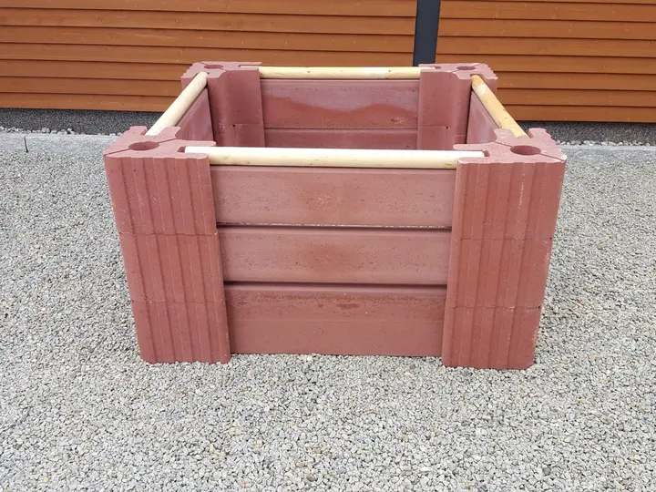 Quadratischer Komposter aus roten Vablo Ecksteinen in drei Lagen, an jeder Seite Betonriegel mit Luftschlitzen und ein helles Rundholz obenauf, auf Kies