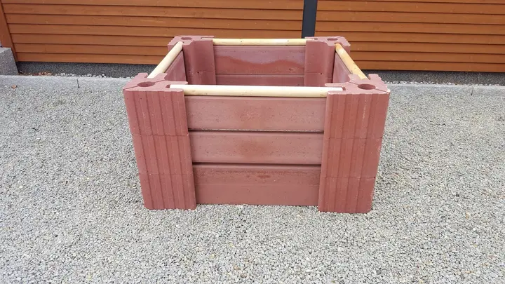 Quadratischer Komposter aus roten Vablo Ecksteinen in drei Lagen, an jeder Seite Betonriegel mit Luftschlitzen und ein helles Rundholz obenauf, auf Kies