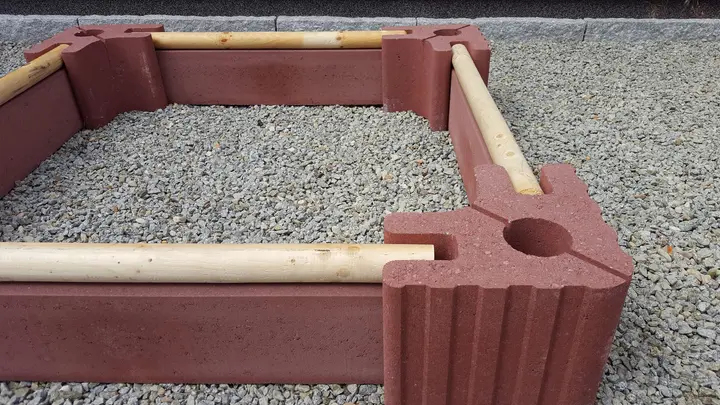 Ecke einer niedrigen Beeteinfassung: roter Vablo Eckstein mit Betonriegel und hellem Rundholz als oberem Abschluss