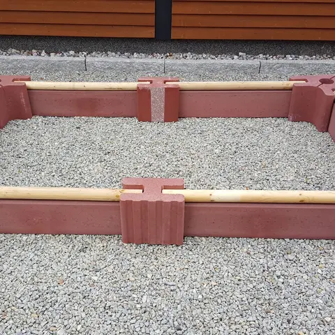 Niedrige längliche Beeteinfassung aus roten Vablo Ecksteinen, Mittelsteinen und Betonriegeln mit hellem Rundholz als oberem Abschluss