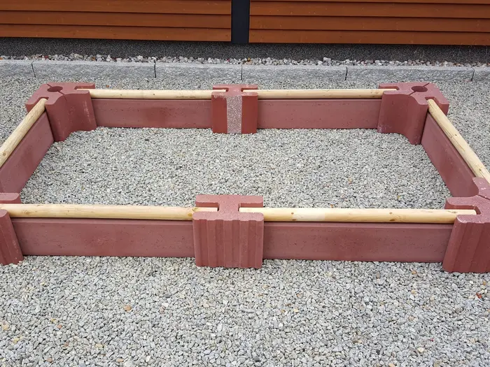 Niedrige längliche Beeteinfassung aus roten Vablo Ecksteinen, Mittelsteinen und Betonriegeln mit hellem Rundholz als oberem Abschluss