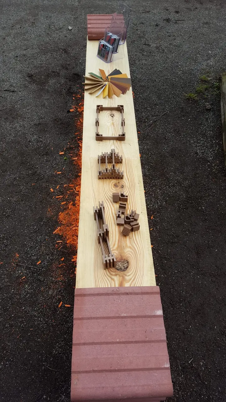 Blick entlang einer langen hellen Holzbohle auf roten Vablo Steinen, mit kleinen Modellen auf der Sitzfläche