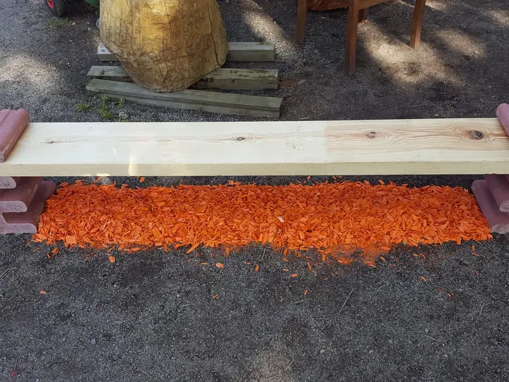 Bank aus einer hellen Holzbohle, die an beiden Enden in liegenden roten Vablo Steinen steckt, darunter orangefarbener Rindenmulch