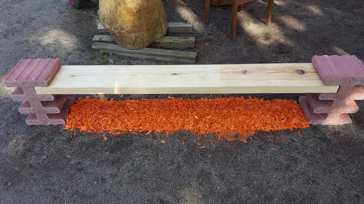 Bank aus einer hellen Holzbohle, die an beiden Enden in liegenden roten Vablo Steinen steckt, darunter orangefarbener Rindenmulch