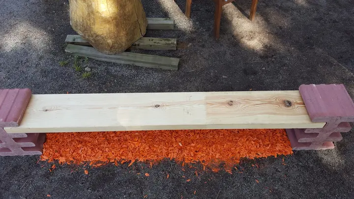 Schrägansicht einer Bank: helle Holzbohle auf zwei liegenden roten Vablo Steinen, im Hintergrund eine Holzskulptur