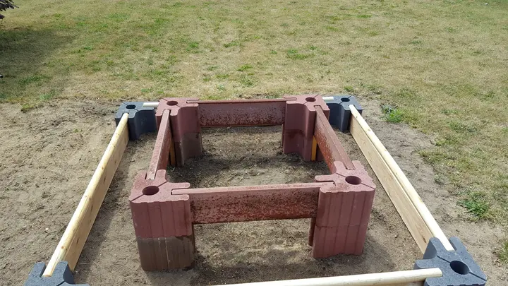 Aufbau eines Stufenbeets auf Sand: innen ein Rahmen aus gestapelten roten Vablo Ecksteinen mit Betonriegeln, außen ein niedriger Rahmen aus anthrazitfarbenen Ecksteinen mit Holzriegeln