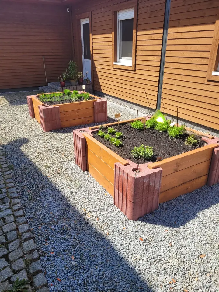 Zwei Hochbeete aus roten Vablo-Ecksteinen und Holzriegeln auf Kies vor einer Holzfassade, frisch bepflanzt.