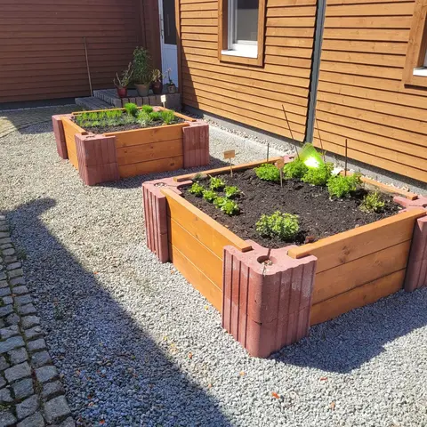 Zwei Hochbeete aus roten Vablo-Ecksteinen und Holzriegeln auf Kies vor einer Holzfassade, frisch bepflanzt.