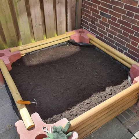 Quadratisches Beet aus roten Vablo-Ecksteinen und Rundholzriegeln in einer Hofecke, frisch mit Erde gefüllt; auf einem Eckstein liegen Arbeitshandschuhe.