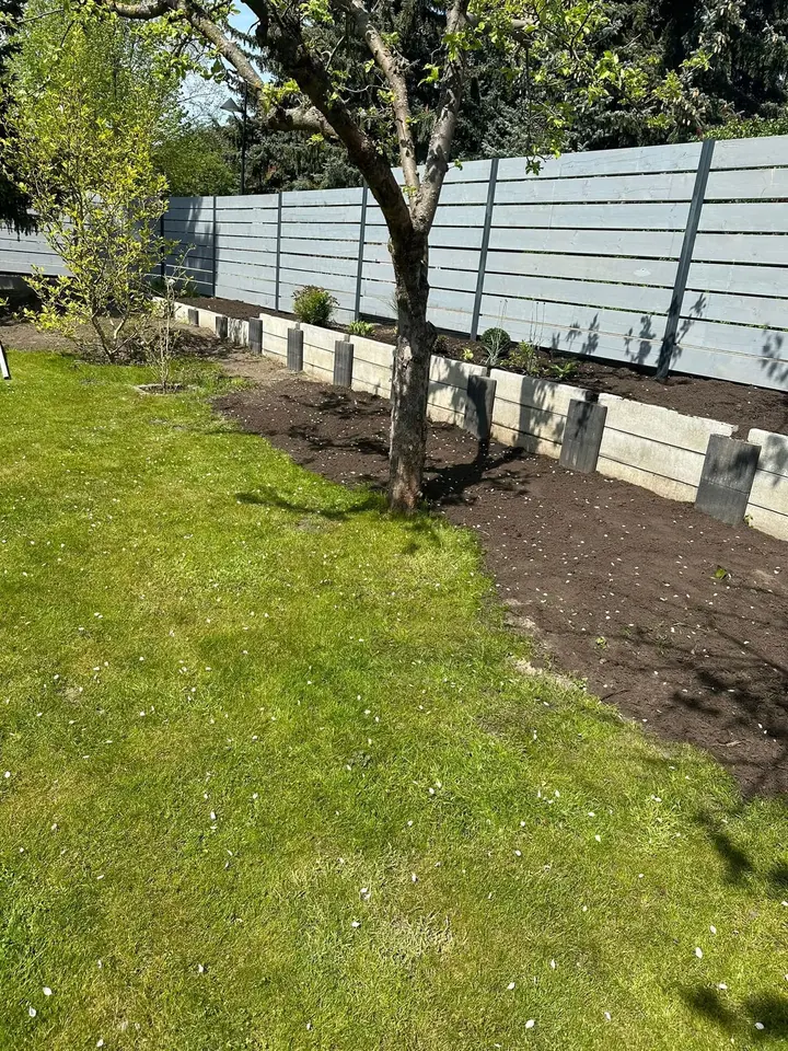 Niedrige Mauer aus anthrazitfarbenen Vablo-Steinen und hellgrauen Betonriegeln entlang eines Holzzauns; dahinter frisch aufgefüllte Erde, davor Rasen.