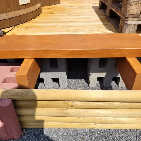Holzterrasse mit offenem Vorderteil: Unter dem Dielenbelag sind Holzbalken und graue Profilsteine zu sehen; vorne begrenzen Rundhölzer zwischen roten Pfosten den Aufbau.
