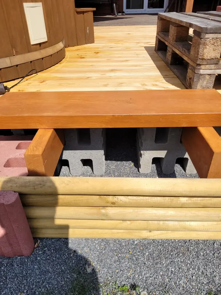 Holzterrasse mit offenem Vorderteil: Unter dem Dielenbelag sind Holzbalken und graue Profilsteine zu sehen; vorne begrenzen Rundhölzer zwischen roten Pfosten den Aufbau.